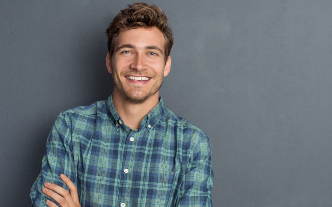 Young handsome man leaning against grey wall with arms crossed. Cheerful man laughing and looking at camera with a big grin. Portrait of a happy young man standing with crossed arms over grey background.